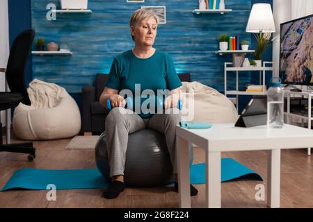 Senior woman exercising body muscles doing arms exercise sitting on swiss ball in living room. Pensioner watching online aerobics training on tablet exercising muscular resistance holding dumbbell Stock Photo