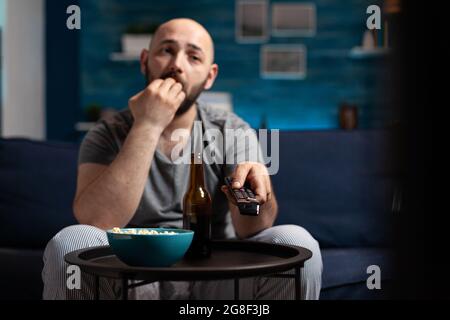 Scared shocked man looking at horror thriller movie at tv, eating popcorn trembled with fright sitting on comfortable sofa. Concentrated alone male watching tv late night enjoying free time Stock Photo