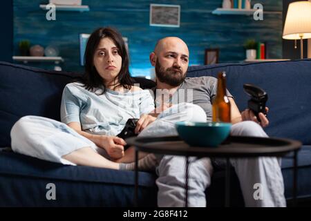 Furious nervous couple losing online videogames championship using joystick playing video games on TV. Frustrated disappointed people arguing because of soccer gaming over late night. Stock Photo