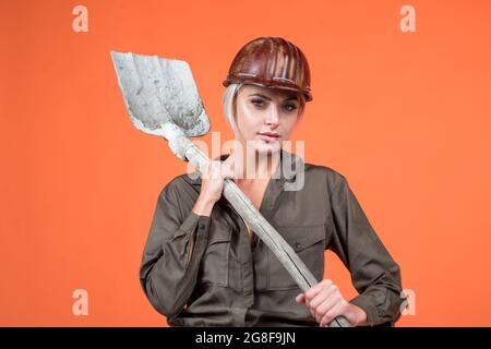 woman hard worker in protective helmet and boilersuit hold shovel on orange background, 1 may Stock Photo