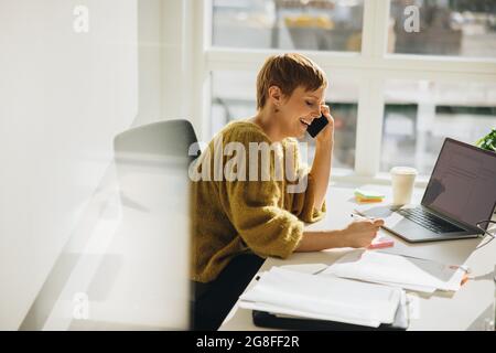 Smiling businesswoman making notes while talking on phone. Female executive working at her desk. Stock Photo
