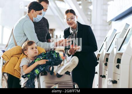 Couple showing mobile phone to flight attendant at airport with son sitting on luggage trolley. Ground attendant assisting traveler family at self ser Stock Photo