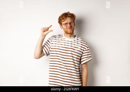 Disappointed young man with red hair and beard, wearing glasses and showing small size, little thing, standing over white background Stock Photo