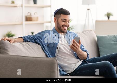 Smiling jew man using cellphone sitting on comfy couch Stock Photo