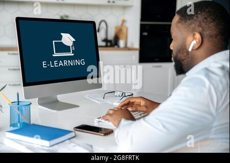 Online education, e-learning concept. African American smart confident guy sitting at home at work desk, uses computer, on computer screen the inscription e-learning. Study remotely Stock Photo