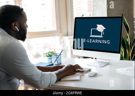 Remote education, e-learning concept. African American smart confident guy sitting at home at work desk, uses computer, on computer screen the inscription e-learning. Knowledges Stock Photo