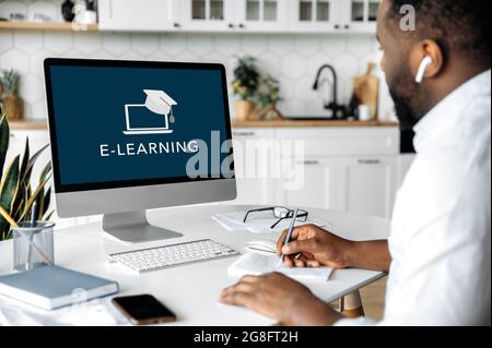 E-learning concept. African American smart confident guy sitting at home at work desk, uses computer, on computer screen the inscription e-learning. Study remotely Stock Photo