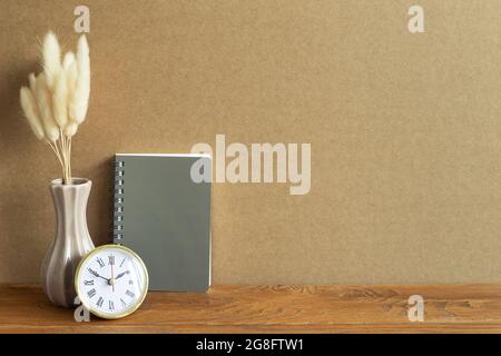 Notebook, clock, dry flower on wooden desk. brown wall background. Workspace Stock Photo