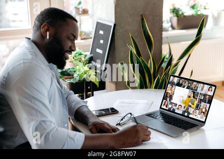 Online virtual meeting, e-learning. Focused african american man, student, sits at workplace, makes notes during online lesson, uses laptop, female teacher and students are on a screen Stock Photo