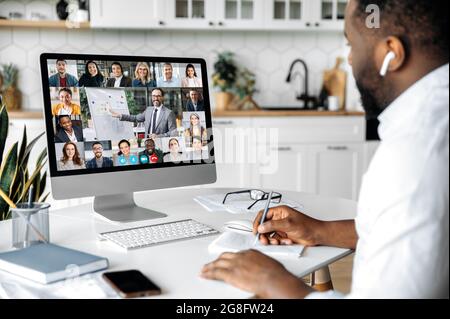 Online education, business seminar. African American smart confident man sit at home at work desk, uses computer, listening to an online financial lecture, taking notes, diverse people on the screen Stock Photo