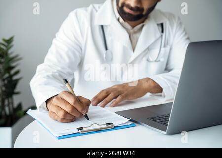 Close up of unrecognizable doctor taking notes sitting at desk front of laptop Stock Photo
