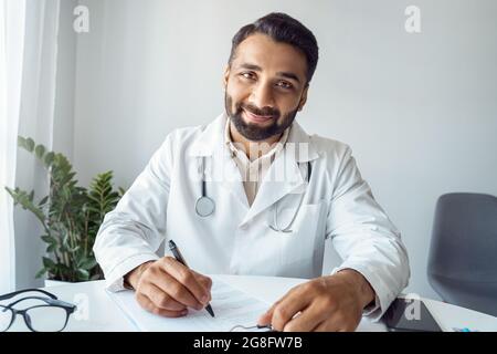 Headshot webcam portrait of smiling indian man doctor writing prescription Stock Photo