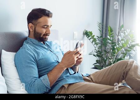 Handsome young adult indian man using smartphone while lying on sofa at home Stock Photo