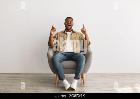 Black Guy Pointing Fingers Upward Sitting Over Gray Wall Stock Photo