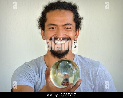 Handsome young Hispanic Latino man with nose ring holds a transparent crystal ball with his mirror image in his right hand and smiles at the viewer. Stock Photo