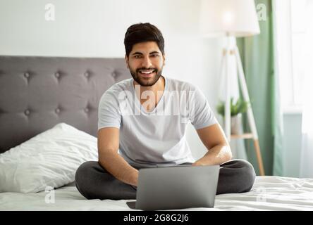 Freelance career. Happy arab man sitting on bed and using laptop computer, working remotely from home Stock Photo