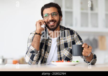 Cheerful arab guy having lunch at home, talking on phone Stock Photo