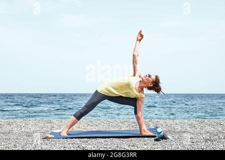 Slim Woman doing Parivritta Parsvakonasana or Revolved Side Angle Pose on the beach in the morning. Strengthening muscles of legs and coordination of Stock Photo