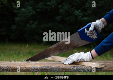 Close up of carpenter wearing gloves and hand sawing wooden plank with old blunt saw in the garden. Stock Photo