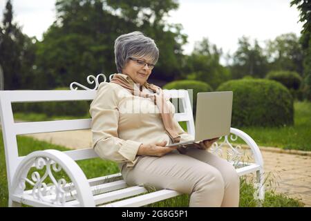 Happy senior lady using laptop sitting on bench relaxing in park outdoors Stock Photo