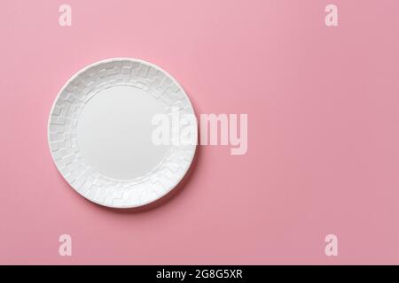 White round ceramic plate with checkered ornament on the edge over textured pastel pink background. Modern empty crockery for stylish food design. Stock Photo