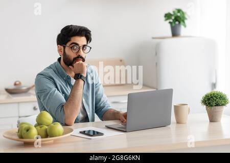 Thoughtful arab businessman thinking of online project, sitting with laptop at kitchen and looking at camera, free space Stock Photo