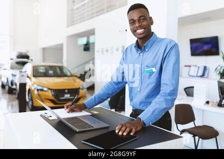 Portrait of happy car salesman standing at work desk, smiling at camera, using laptop and touch pad in auto dealership Stock Photo