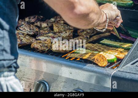 Close view of male hand handling meat and vegetables with tongs on the hot grill of a barbecue set Stock Photo