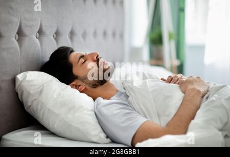 Deep male sleep. Young arab guy sleeping peacefully in his comfortable bed at home, lying with closed eyes on back Stock Photo