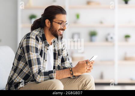 Positive long-haired middle-eastern guy sitting on couch, using cellphone Stock Photo