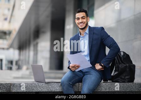 Handsome arab entrepreneur sitting by office building with laptop Stock Photo