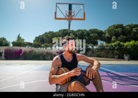 tired basketball player after the match, sits on the basketball field with bottle of water. fluid and water recovery and hydration after training Stock Photo