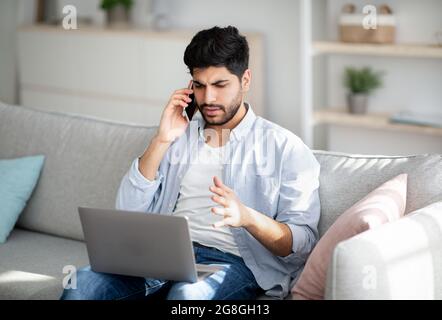 Computer problems. Displeased arab man talking on smartphone and looking on laptop screen at home Stock Photo
