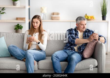 Mature couple using mobile devices, sitting apart on sofa, not paying attention to each other, playing online games Stock Photo