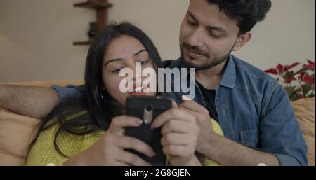 Young Indian couple happily chatting while sitting on the sofa and using the phone Stock Photo