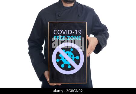 Covid free zone sign. Cook in black uniform on white background holding a board, COVID-19 free zone text . Restaurant disinfected areas, vaccinated on Stock Photo
