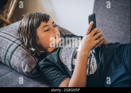 Teen boy checking social media holding smartphone, using mobile phone app playing game, relaxing on sofa at home. Emotional isolation and depression Stock Photo