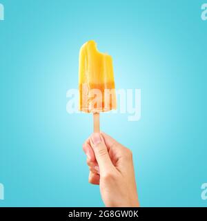 Woman holds bitten ice cream on blue background with copy space. Color frozen fruit popsicle. Stock Photo