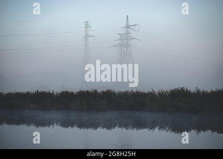 A power line passes over the river in Latvia. Morning shot in the haze on the background of the forest. Lots of pylons in meadow looks like big giants Stock Photo
