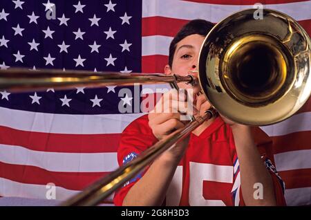 Young boy in bedroom with an American flag on his wall playing the trombone looking at camera Stock Photo