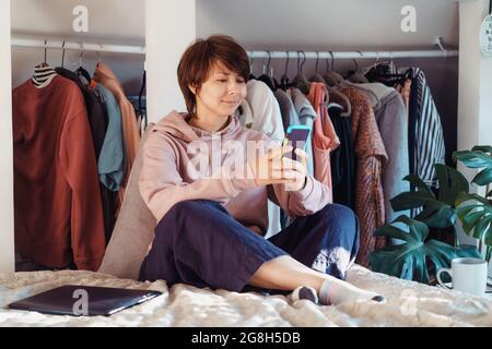 Woman sitting on the bed and using her smartphone in an apartment with an open wardrobe. Cozy home. Online communication. Remote work. Scrolling Stock Photo