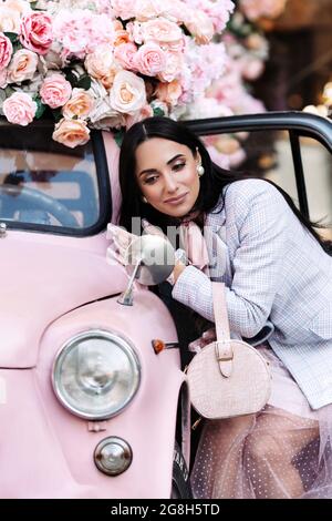 Portrait of stylish elegant female model in pink outfit look in mirror of small retro car outdoors Stock Photo
