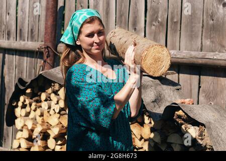 country woman peasant in traditional dress and headscarf carries a log from a woodpile Stock Photo