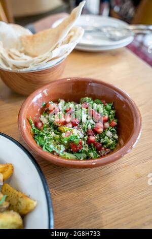 Close-up of a tasty tabbouleh salad with pomegranate seeds in a brown ceramic bowl, plates in the background on a wooden table Stock Photo