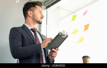 Portrait of concentrated male with eyeglasses writing on notebook while looking at sticky notes on glass wall in office. Businessman developing plan Stock Photo