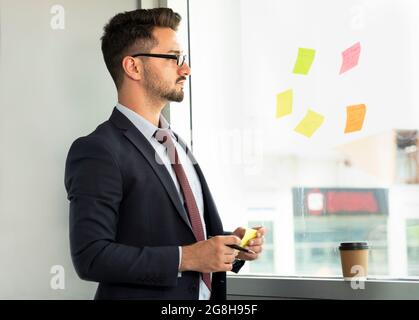 Portrait of concentrated male executive with eyeglasses looking at sticky notes on glass wall in office. Businessman focused on business activity Stock Photo