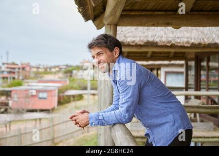 Young man in a light blue shirt leaning on the wooden railing of the terrace of his cabin while smiling. Stock Photo