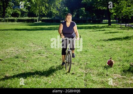 Young healthy woman rides bike in park with small dog running near. Stock Photo