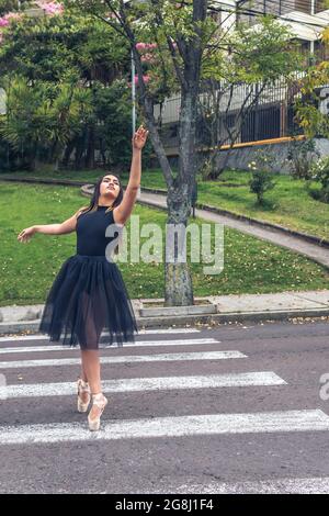 Ballerina in black suit and ballet shoes dancing in the street in a pedestrian crossing with one arm up Stock Photo