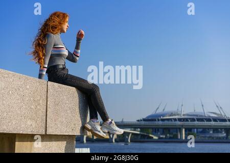 Beautiful young woman with long hair of Caucasian nationality , in casual clothes sits on the embankment of the river on a spring sunny day with a view of the bridge Stock Photo